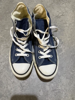 Converse All Star blå high tops - Säljer ett par klassiska Converse All Star high tops i marinblått med vita snören och gummisula. Skorna har rund tå, vit tåhätta och tydliga vita sömmar. Märket syns på sidan och bak på hälen. Materialet är textil med gummisula.