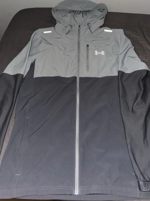 Grå och svart vindjacka Under Armour - Snygg tvåfärgad vindjacka från Under Armour i grått och svart med huva och hel dragkedja framtill. Jackan har en bröstficka med dragkedja och logga på bröstet. Perfekt för utomhusaktiviteter och träning när det blåser.