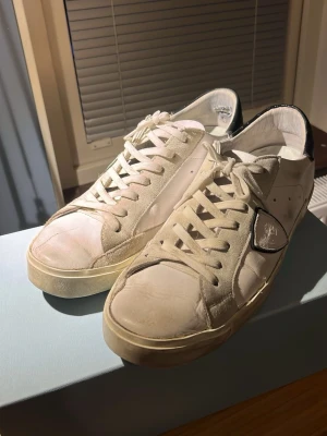 Philippe Model vita sneakers herr 44 - Säljer ett par vita Philippe Model sneakers med svarta detaljer på hälen och signaturmärket på sidan. Skorna har snörning, rund tå och platt sula. Materialet är en mix av skinn och mocka, vilket ger en schysst kontrast. Perfekt för dig som gillar stilrena och klassiska sneakers. Allt OG ingår dvs dustbag och låda. 