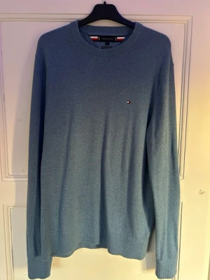 Blå stickad tröja Tommy Hilfiger  - Snygg blå stickad tröja från Tommy Hilfiger i storlek L. Tröjan har rund hals, ribbade muddar och ett litet broderat Hilfiger-logo på bröstet. Materialet är en mjuk blandning av pima bomull och kashmir som känns lyxigt mot huden. Perfekt för dig som gillar stilrena plagg.