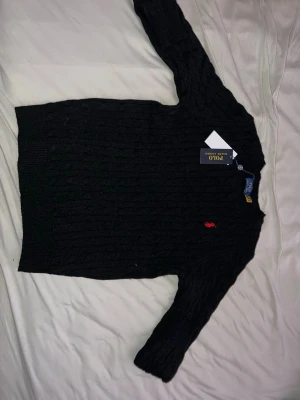 Svart stickad tröja Polo Ralph Lauren - Svart kabelstickad tröja från Polo Ralph Lauren i storlek S. Klassisk design med rund hals och rött broderat logomärke på bröstet. Långärmad och ribbad vid muddar och nederkant. Perfekt för dig som gillar stilrena och tidlösa plagg.
