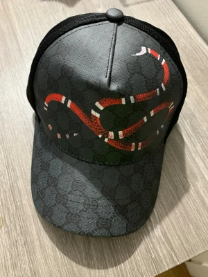 Svart G keps - Säljer en svart keps från med ett diskret GG-mönster och en stor röd och vit orm på framsidan. Kepsen har mesh på baksidan och justerbar rem med blå och röd detalj. Insidan är märkt med -logga. Perfekt för dig som vill sticka ut.