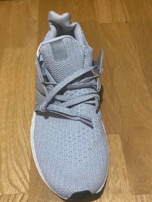 Adidas Ultraboost grå sneakers - Säljer ett par Adidas Ultraboost sneakers i ljusgrå färg med stickad ovandel och klassiska tre ränder på sidan. Skorna har snörning, platt sula och rund tå. Perfekta för dig som gillar stilrena och bekväma sneakers med modern design.