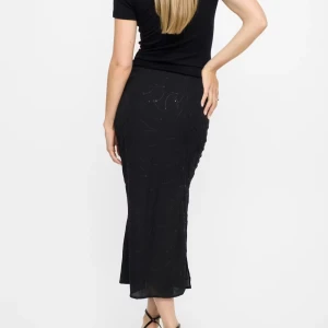 Svart långkjol med paljettdetaljer - Säljer en svart långkjol med glittriga paljettdetaljer och subtilt mönster. Kjolen har en slits på sidan som ger extra edge och är figurnära med lätt utsvängd nederdel. Perfekt för dig som gillar att sticka ut med stilrena detaljer.