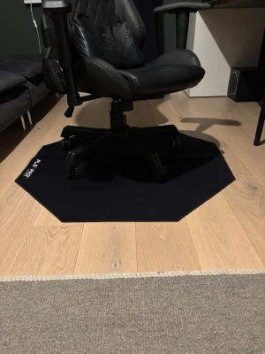 Svart Florpad gamingmatta - Säljer en svart Florpad gamingmatta med oktagonformad design och vit logga i hörnet. Mattan är tillverkad i slitstark syntet och passar perfekt under gamingstolen för att skydda golvet och ge extra komfort. Stilren och enkel att matcha med ditt setup. Som skyddar golvet kan användas till kontor eller andra slags stolar