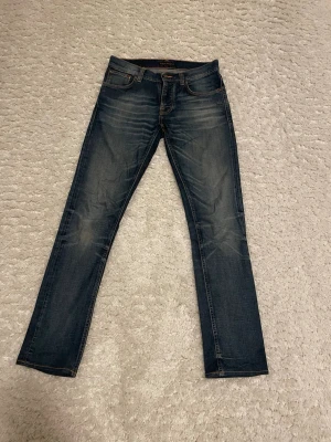 Nudie Grim Tim  - Snygga mörkblå jeans från Nudie med klassisk femficksdesign och kontrastsömmar. Jeansen har modellen Grin Tim och är tillverkade i slitstarkt denim med lätt tvättade detaljer på framsidan och baksidan, Jeansen har storleken 30W och 34L. Perfekt för dig som behöver ett par nya jeans.