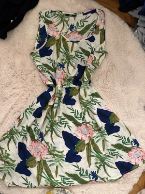 Blommig sommarklänning från ONLY - Snygg kort klänning från ONLY med blommigt mönster i blått, rosa och grönt på vit botten. Ärmlös modell med rund hals och markerad midja. Perfekt för varma dagar och har en lätt och luftig känsla.