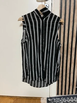 Svartvit randig ärmlös skjorta - Snygg ärmlös skjorta med svart botten och vita vertikala ränder. Skjortan har en hög krage och knappar framtill. Materialet är lätt och luftigt, perfekt för varmare dagar. Passformen är normal och skjortan har en modern, stilren look.