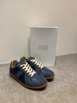 Maison Margiela blå sneakers - PRIS KAN DISKUTERAS🔥🔥🔥.        Maison Margiela sneakers i marinblått med beige snörning och gummisula. Skorna har en mix av mocka och skinn på ovandelen, rund tå och platt sula. Klassisk design med diskreta detaljer och logotyp under sulan. Perfekt för dig som gillar stilrena och exklusiva sneakers.