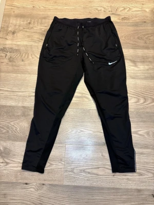 Nike Running - Svarta mjukisbyxor från Nike med smal passform och dragsko i midjan. Byxorna har dragkedjeförsedda fickor och en vit Nike-logga på låret. Tillverkade i polyester, perfekta för träning eller chill. Snygga detaljer med text på snörena.
