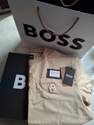 Beige kortärmad t-shirt från BOSS - Ny beige t-shirt från BOSS med broderad logga på bröstet. Klassisk rund hals och korta ärmar. (Bred i modellen). Tillverkad i mjuk bomull. Perfekt för dig som gillar stilrena och exklusiva plagg med diskreta detaljer.