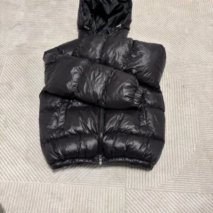 Svart pufferjacka med huva - Säljer en svart pufferjacka med luva och dragkedja framtill. Jackan har en loose passform och är tillverkad i glansig syntet med vadderade sektioner. Perfekt för kalla dagar och har långa ärmar med elastiska muddar för extra komfort.