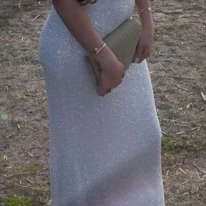 Glittrig långklänning med öppen rygg - Supersnygg långklänning i vitt med glittrigt tyg och smala axelband. Klänningen har öppen rygg med snörning och en figurnära passform som ger en elegant siluett. Perfekt för dig som vill sticka ut och glänsa på nästa stora tillställning!