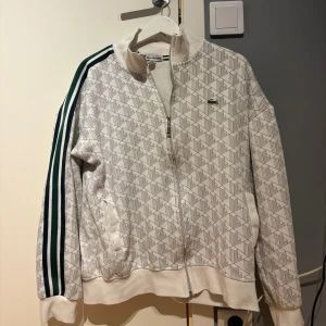 Vit mönstrad zip Hoddie från Lacoste - Vit zip Hoddie från Lacoste med geometriskt mönster i grått och gröna ränder längs ärmarna. Jackan har dragkedja framtill, ståkrage och muddar vid ärmslut och midja. Klassisk krokodillogga på bröstet och två fickor framtill.