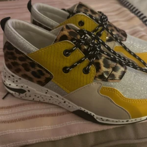 Färgglada sneakers med leopardmönster - Unika sneakers från Justin Shoes i storlek 41 med cool mix av gult, silver, beige och svart samt detaljer i leopardmönster. Skorna har snörning, vit sula med svarta prickar och glittriga partier. Perfekta för dig som vill sticka ut med din stil.