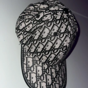 Christian Dior keps hel äkta - Snygg keps från Christian Dior med ikoniskt monogrammönster i svart och silver. Kepsen har böjd skärm med svart kant och justerbar passform. Materialet är polyester med detaljer i syntet. Perfekt för dig som vill sticka ut med en lyxig accessoar.