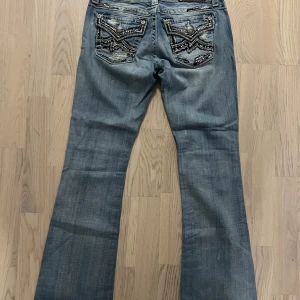 Miss Me bootcut jeans med detaljer - Snygga ljusblå bootcut jeans från Miss Me med coola broderade bakfickor och nitar. Jeansen har slitna detaljer, silverfärgade knappar och kontrastsömmar. Perfekt för dig som vill sticka ut med unika detaljer och en klassisk bootcut passform.