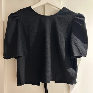 Svart blus med knytning från Dazy - Svart blus från Dazy i storlek XL med korta puffärmar och snygg struktur i tyget. Baksidan har flera knytband som ger en cool och unik look. Blusen är tillverkad i ett lätt och något stretchigt material som känns bekvämt mot huden. Passar mig som är S/M