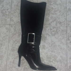 Svarta stövlar med klack och spänne - Snygga mörka (ser svarta ut men är lite mörk brun/röda)  stövlar i skinn med hög klack och spetsig tå. Köpta i Spanien (bor här), för 70 euro. Använd två gånger! Storlek 38!