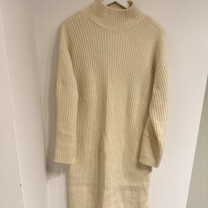 Beige stickad klänning New Look - Mysig beige stickad klänning från New Look i ribbat material. Klänningen har polokrage, långa ärmar och slitsar i sidorna. Perfekt för dig som gillar en enkel och stilren look.