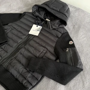 Moncler cardigan  - Svart cardigan från Moncler med huva, ribbstickade ärmar och detaljer. Jackan har dragkedja framtill, sidofickor och Moncler-logga på ärmen. Materialmix med stickad insida och quiltad utsida ger en schysst look. Perfekt för dig som gillar street och premium.