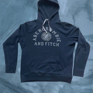 Mörkblå hoodie från Abercrombie & Fitch - Mörkblå hoodie från Abercrombie & Fitch med broderad logga och text framtill. Tröjan har huva med snörning, känguruficka och långa ärmar. Perfekt för en chill och avslappnad stil nu till hösten/vintern. Pris kan diskuteras vid snabb affär! Ha det gött// Gidlöfs