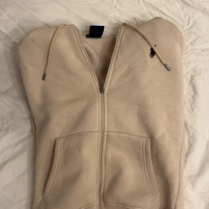 Beige hoodie från Polo Ralph Lauren - Snygg beige hoodie från Polo Ralph Lauren med dragkedja, huva och snörning. Tröjan har broderad svart logga på bröstet, två fickor fram och är tillverkad i mjuk bomull. Perfekt för chill dagar och enkel att matcha med jeans eller joggers.
