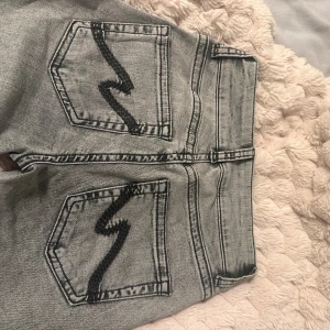 Grå jeans - Säljer ett par grå jeans från Nelly med coola broderade detaljer i svart på bakfickorna. Jeansen har låg midja, klassisk femficksdesign och dragkedja med dubbla knappar framtill. Perfekt för dig som gillar en avslappnad och trendig stil.