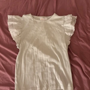 Ljusgrön ribbad topp med volangärmar - Supersöt ljusgrön topp med ribbad struktur och breda volangärmar i broderad bomull. Ärmlös modell med rund halsringning och romantiska detaljer på ärmarna som ger en extra touch. 