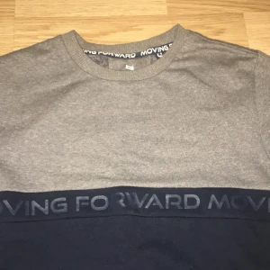 Grå och mörkblå tröja Moving Forward - Snygg tvåfärgad tröja från Moving Forward i grått och mörkblått. Tröjan har rund hals och ett band med texten 'MOVING FORWARD' över bröstet. Tillverkad i mjuk bomull, perfekt för chill dagar. Långärmad och med normal passform.