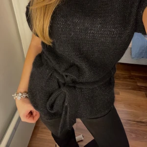 Svart stickad tröja med knyt - Jättesnygg svart stickad tröja från Green House i storlek M. Modellen har korta ärmar, rund hals och ett snyggt knyt i midjan. Perfekt att styla! 35% mohair och 10% ull