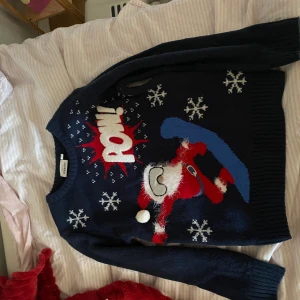 Mörkblå jultröja - Rolig mörkblå stickad jultröja från Kappahl!! med ett stort motiv av en snowboardande tomte, snöflingor och texten 'POW!' i vitt och rött. Tröjan har lång ärm, ribbade muddar och fluffiga detaljer på tomtens skägg och mössa. Storlek 134/140 barn!