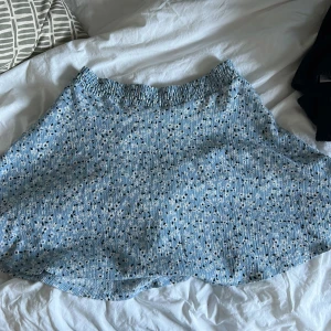 Blå prickig kortkjol från 157 - Säljer en blå kortkjol från 157 i storlek XS/S. Kjolen har ett blommigt mönster i svart och vitt, med en ribbad och elastisk midja. Materialet är mjukt och känns som bomull, perfekt för dig som gillar bekväma och snygga plagg med lite struktur.