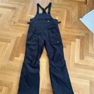 Svarta skidbyxor från Dope Snow - Svarta täckbyxor från Dope Snow med hängslen och flera fickor framtill. Byxorna har justerbara knappar i midjan och är tillverkade i slitstarkt syntetmaterial, perfekta för vinterns äventyr. Snygg logga på bröstet och cool design.