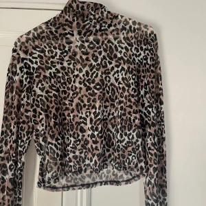 Leopardmönstrad mesh polotopp - Säljer en transparent polotopp i mesh med leopardmönster i svart, beige och brunt. Toppen har lång ärm och croppad passform
