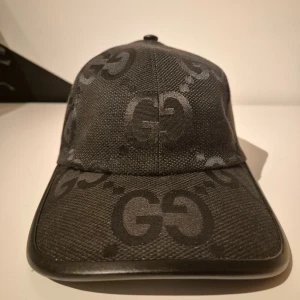 Svart keps med GG-mönster - Snygg svart kepsi med klassiskt GG-mönster över hela kepsen. Justerbar rem baktill i skinn och böjd skärm. Tillverkad i slitstarkt material med exklusiv känsla och diskret glans i tyget. Storlek S, 57 cm.
