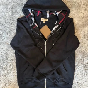 Burberry Zipper - Lyxig hoodie från Burberry i svart med den klassiska rutiga huvan som detalj. Ikonisk och tidlös design med högsta kvalitet – perfekt för både vardag och mer dressade tillfällen.  ♦️ Helt ny – med originaltag kvar ☑️ ⚜️ ☑️ ♦️ Exklusiv Burberry-detalj i huvan ☑️  📦 Snabb leverans & trygg affär 