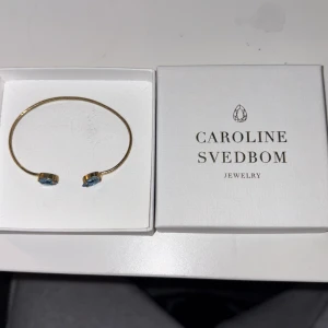 Guldarmband med blå stenar Caroline Svedbom - Snyggt och stilrent guldarmband från Caroline Svedbom med två gnistrande blå stenar i oval form på öppna ändarna. Perfekt accessoar för att lyfta din outfit med lite färg och elegans. Kommer i original ask. Nypris 799