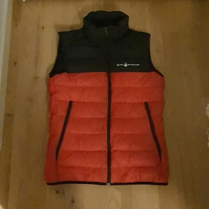 Röd och svart dunväst från Sail Racing - Snygg dunväst från Sail Racing i rött och svart med hög krage och dragkedja framtill. Västen har två fickor med dragkedja. Varm med certifierat dun. Vind- och vattenavvisande material. Snygg väska allmänt bra kvalité inköptes i fjol för 1300 i en stil racing butik strl 160 som ny använts ett par fåtalgånger. Kan gå ner I pris vid snabb äffär.