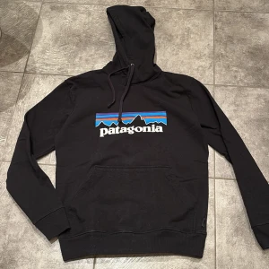 Svart Patagonia hoodie XS - Svart hoodie från Patagonia i storlek XS med klassisk logga och bergmotiv i blått, rött och orange på bröstet. Tröjan har huva med snörning, magficka och är tillverkad av återvunnen polyester och bomull med lite elastan för extra komfort. Nypris 1100