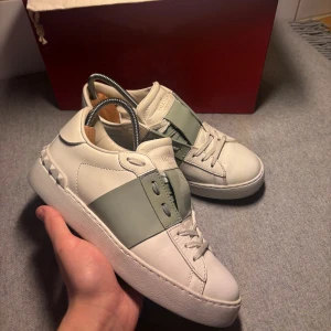 Valentino Garavani Open skor - Säljer ett par Valentino Garavani Open sneakers i vitt skinn med bred grön rem över vristen och ikoniska nitar baktill. Klassisk rund tå, platt sula och snygga detaljer. Perfekta för dig som vill ha en lyxig och stilren sneaker med edge.