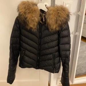 Svart pufferjacka med päls från Hollies - Svart pufferjacka från Hollies med stor fluffig fuskpäls på huvan. Jackan har dragkedja framtill, två fickor med dragkedja och snyggt mönstrat innerfoder. Perfekt för kalla dagar och ger en cool look.