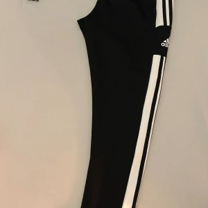 Svarta Adidas träningsbyxor med vita ränder - Svarta träningsbyxor från Adidas med klassiska vita ränder längs sidorna och Adidas-logga på låret. Byxorna har en slim passform och är gjorda i ett mjukt och stretchigt syntetmaterial som passar perfekt för träning eller chill. i storlek M 