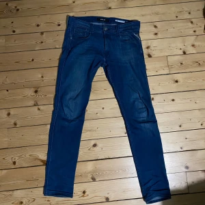 Blå Replay Anbass jeans W32 L34 - Säljer ett par blå Replay Anbass jeans med klassisk femficksdesign och snygga slitningar. Jeansen har smal passform och raka ben, tillverkade i stretchig denim för extra komfort. De har även en snygg slitning vid grenen. Perfekta för dig som gillar en modern och stilren look. Även perfekta nu till hösten!🔥