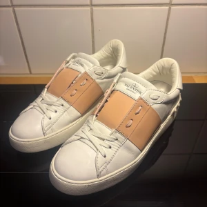 Valentino Garavani Open skor - Snygga Valentino Garavani Open sneakers i vitt skinn med bred ljusrosa panel över snörningen. Klassisk rund tå, platt sula och coola nitar baktill. Perfekta för dig som vill ha en lyxig och trendig look med en touch av färg.