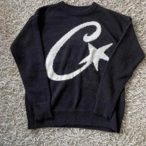 Svart stickad Corteiz tröja - Svart stickad tröja med stor vit C och stjärna på framsidan. Rund hals och långa ärmar, mjuk och skön passform. Perfekt för dig som gillar grafiska detaljer och vill sticka ut lite extra. Passar till jeans eller cargo för en chill look.