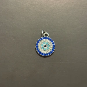 Rund blåvit hängsmycke i silverfärg - Säljer ett runt hängsmycke med glittrande blå och vita stenar i cirkelmönster. Framsidan har flera lager av små stenar i olika blå nyanser och vitt, medan baksidan är silverfärgad med ett strålande mönster. 