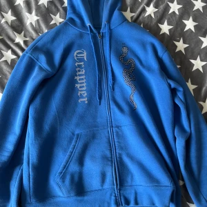 Made by trappers hoodie - Blå hoodie från Trapper med dragkedja, huva och känguruficka. På framsidan finns ett strassmönster med texten 'Trapper' och en cool orm i strass. Mjuk fleece på insidan och snörning i huvan. Perfekt för dig som gillar streetstyle.