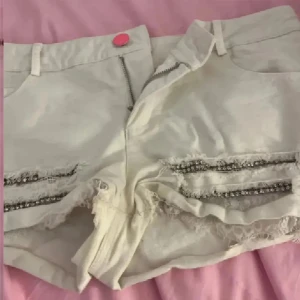 Vita shorts med strass och fransar - Snygga vita shorts med råa fransade kanter och dubbla rader av strassdetaljer framtill. De har en hög midja och en rosa knapp som cool kontrast. Perfekta för sommarens alla äventyr och ger en trendig vibe till din outfit.
