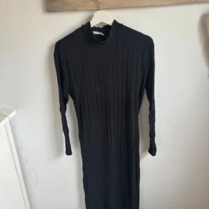 Svart ribbad långklänning från MNG - Svart långklänning från MNG med ribbad struktur och lång ärm. Klänningen har en hög krage och ett avtagbart bälte i midjan för extra form. Perfekt för dig som gillar en stilren och enkel look.